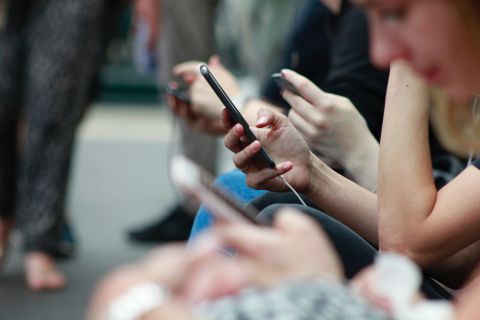 A row of people hold smartphones. It is mainly people’s hands which are visible, as well as the profile of an out-of-focus face which is lowered to look at a phone.